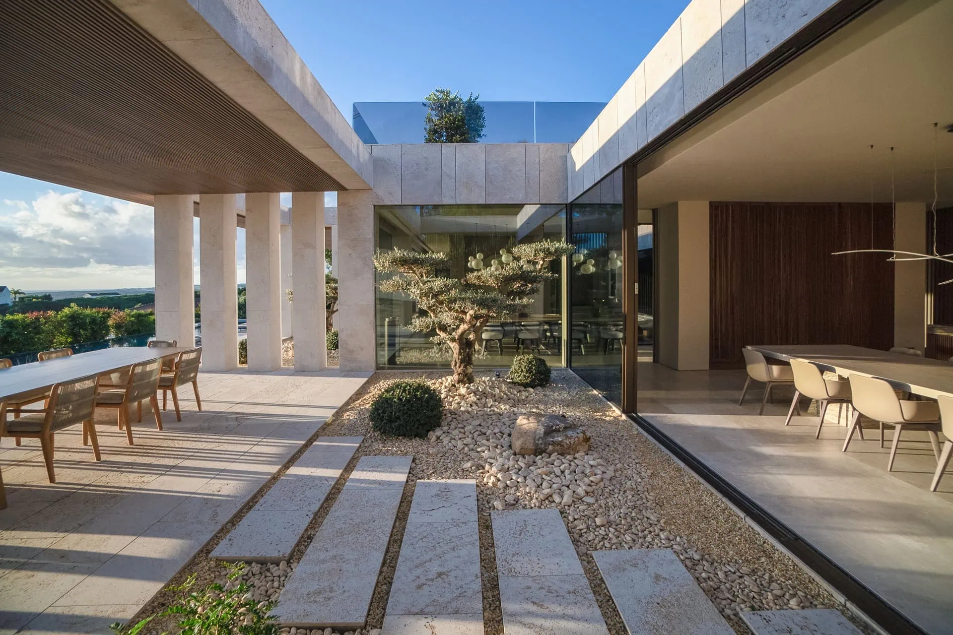 TAI project interior courtyard by ARK Architects: open-plan dining area with stone flooring, gravel garden, bonsai tree, wooden accents, and glass walls blending indoor-outdoor spaces